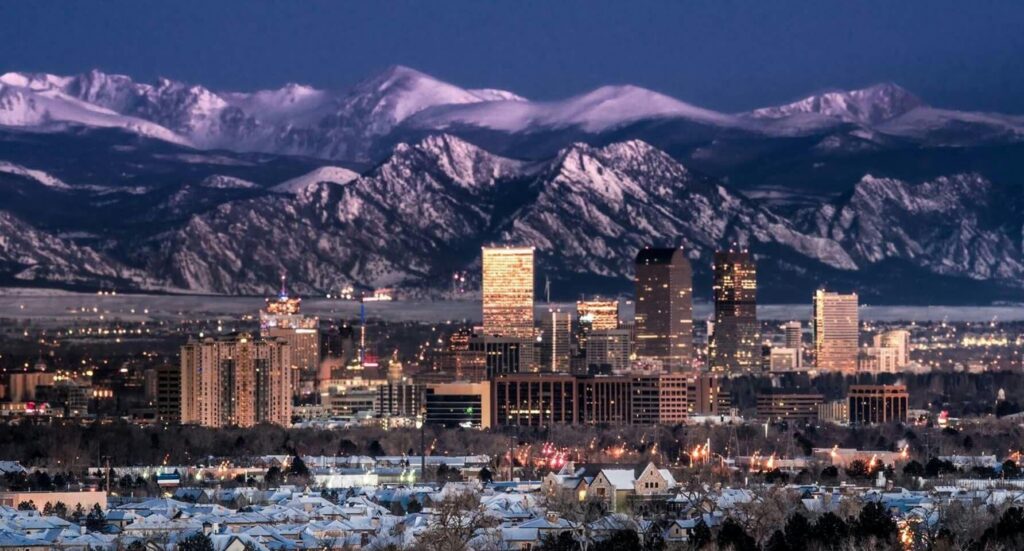 A city with illuminated buildings at dusk, nestled in front of a mountain range with snow-capped peaks.